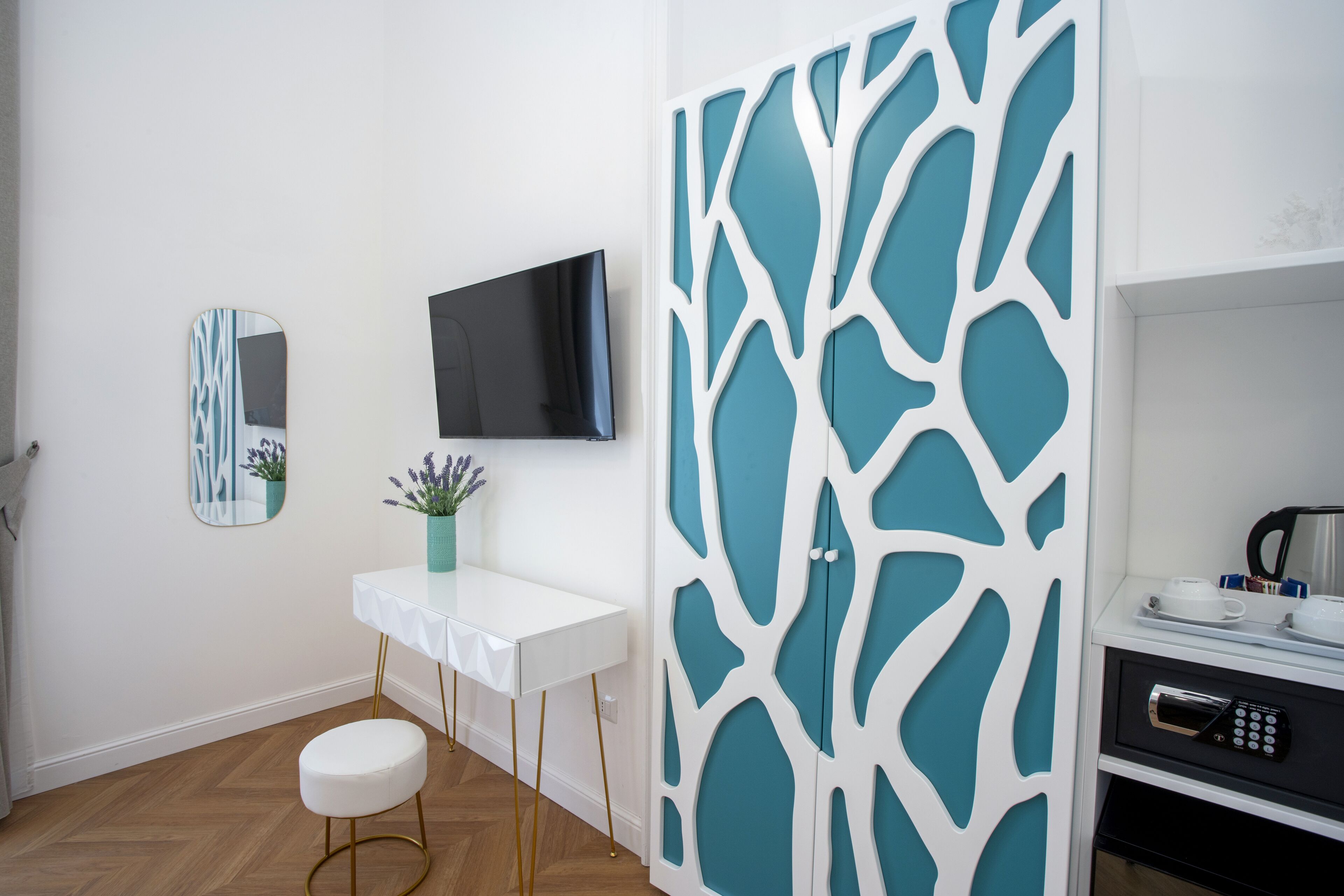 Una habitación moderna con un tocador blanco, un televisor de pantalla plana, un panel decorativo color turquesa y una kitchenette con microondas.