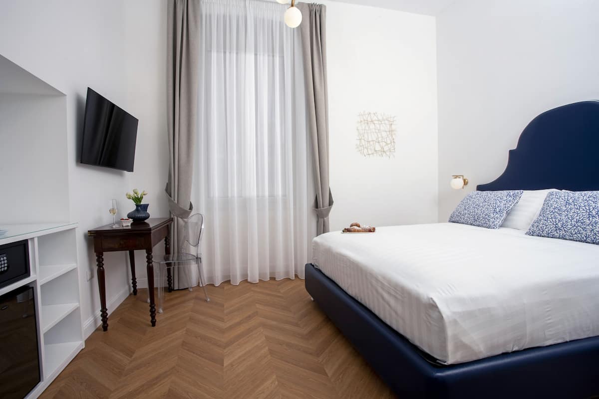 A bedroom with a bed, a TV mounted on the wall, a desk with a chair, and a window with curtains.