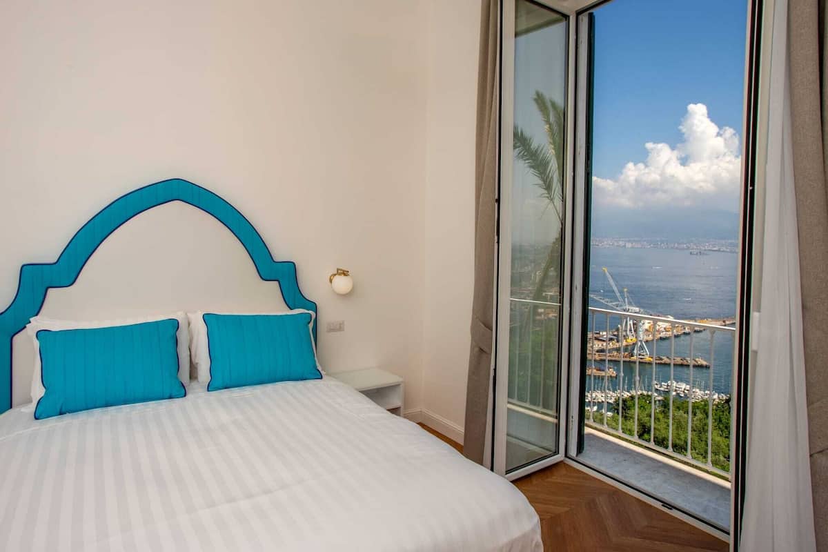 A bedroom with a bed, two blue pillows, a nightstand, and a view of a harbor with boats and a bridge.