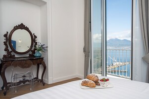 Un dormitorio con un ventanal grande, un aparador de madera, una silla transparente y una bandeja con cruasanes y fresas.