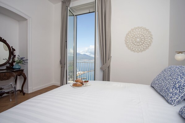 A bedroom with a bed, a dresser, a mirror, a window with a view of a city and mountains, and a decorative wall piece. - Relais al Castello (Castellammare di Stabia)