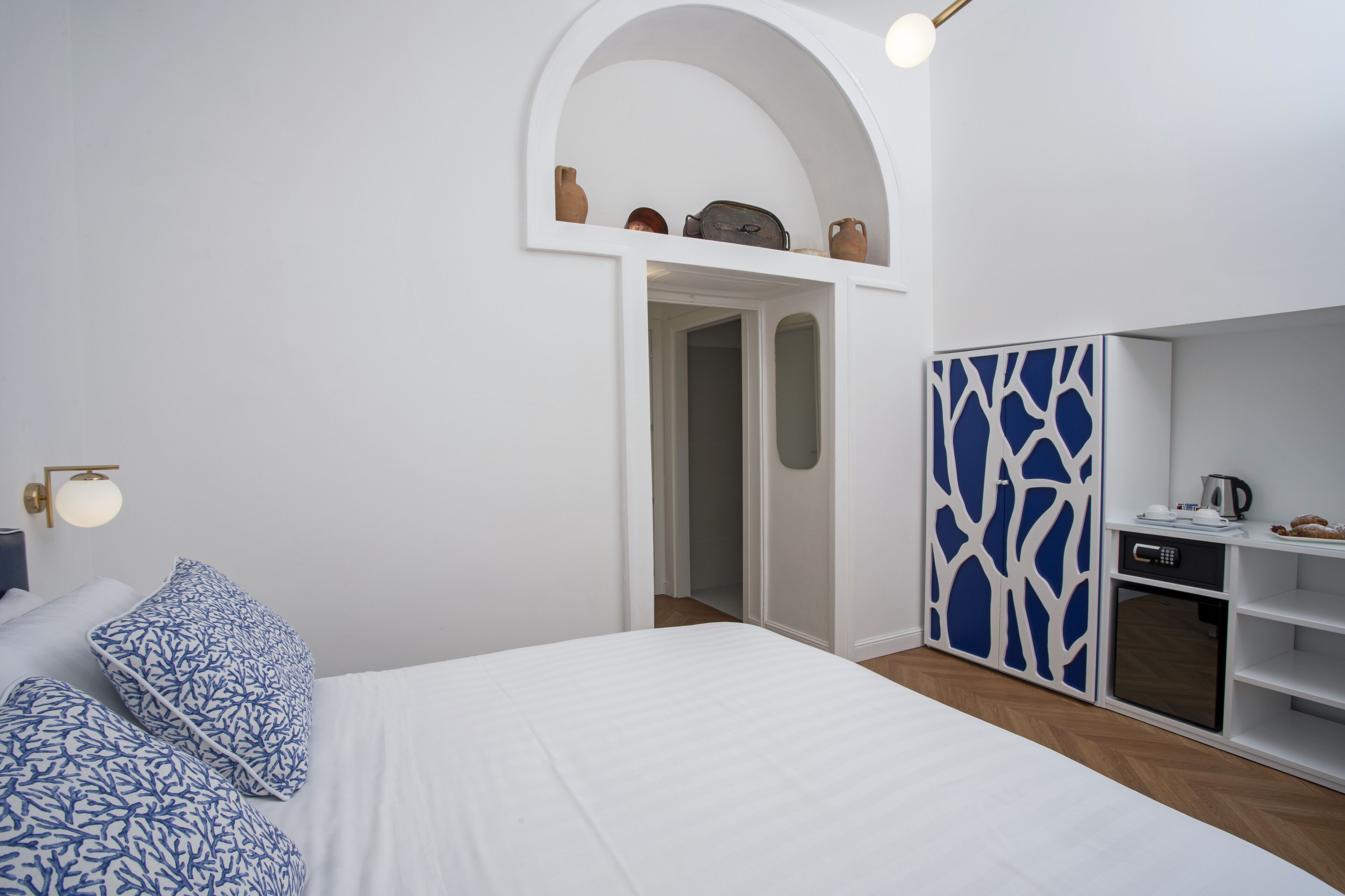A modern bedroom with a bed, blue patterned pillows, a white wardrobe, and a small kitchenette.