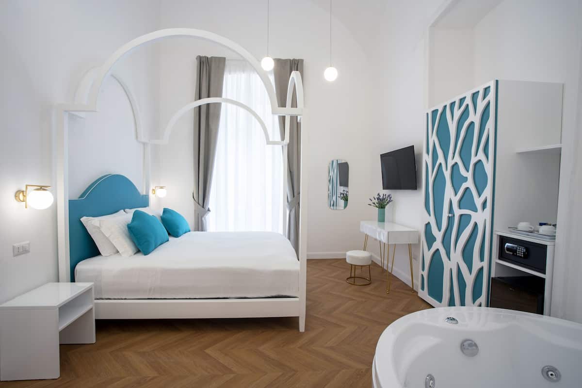 A modern bedroom with a white bed, blue headboard, and a large window with curtains. There is a white nightstand, a small white table, and a white wall-mounted TV.