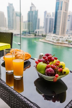 A table with a fruit bowl, glasses of orange juice, and pastries, overlooking a cityscape with buildings and water.