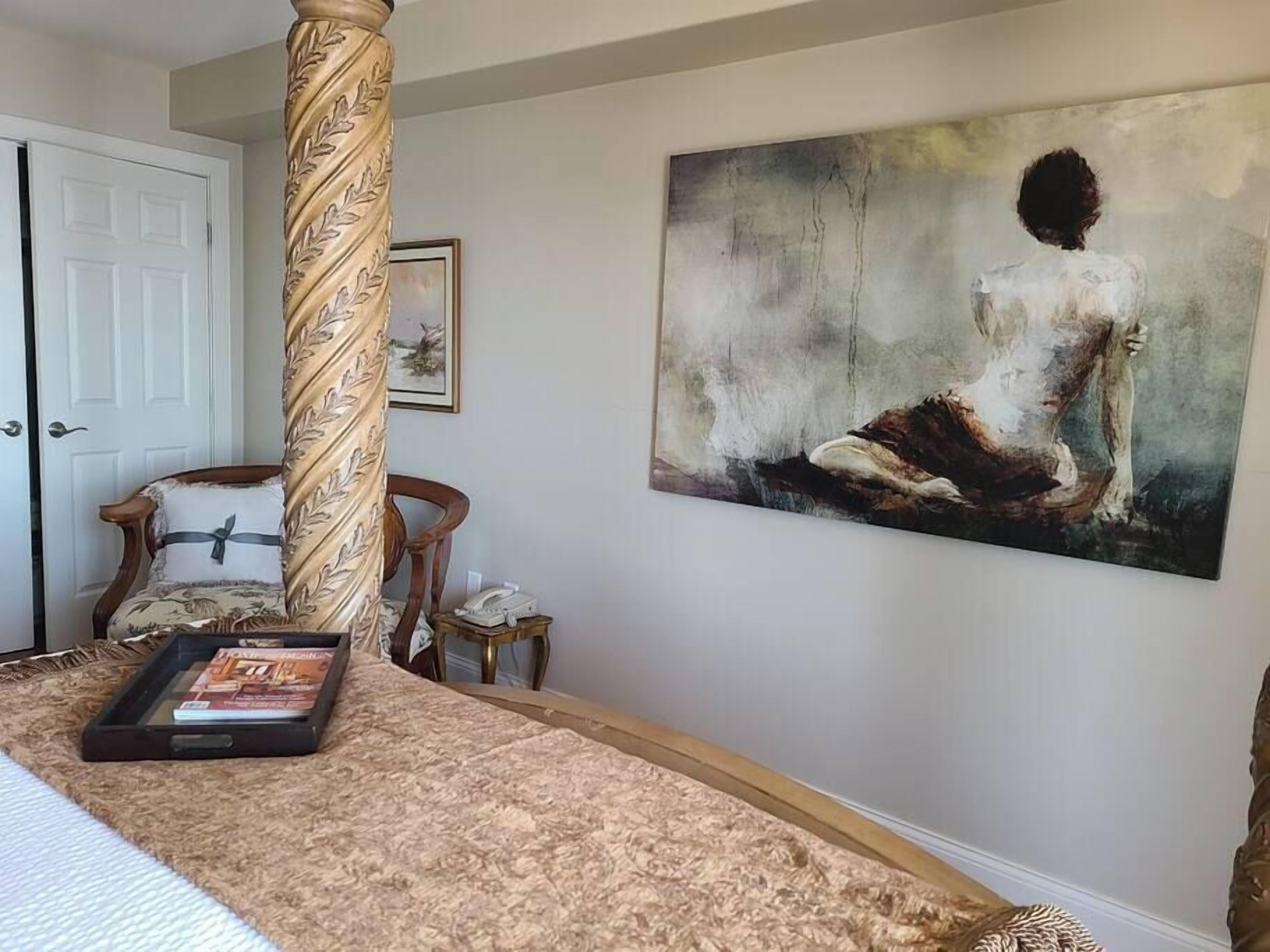 Een slaapkamer met een groot schilderij van een vrouw op een rots, een houten stoel, een nachtkastje met een telefoon en een tijdschrift op het bed.