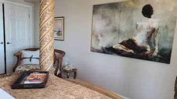 Een slaapkamer met een groot schilderij van een vrouw op een rots, een houten stoel, een nachtkastje met een telefoon en een tijdschrift op het bed.