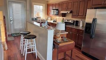 A kitchen with wooden cabinets, a stainless steel refrigerator, a microwave, and a stove. There are two stools and a small table with a lamp.