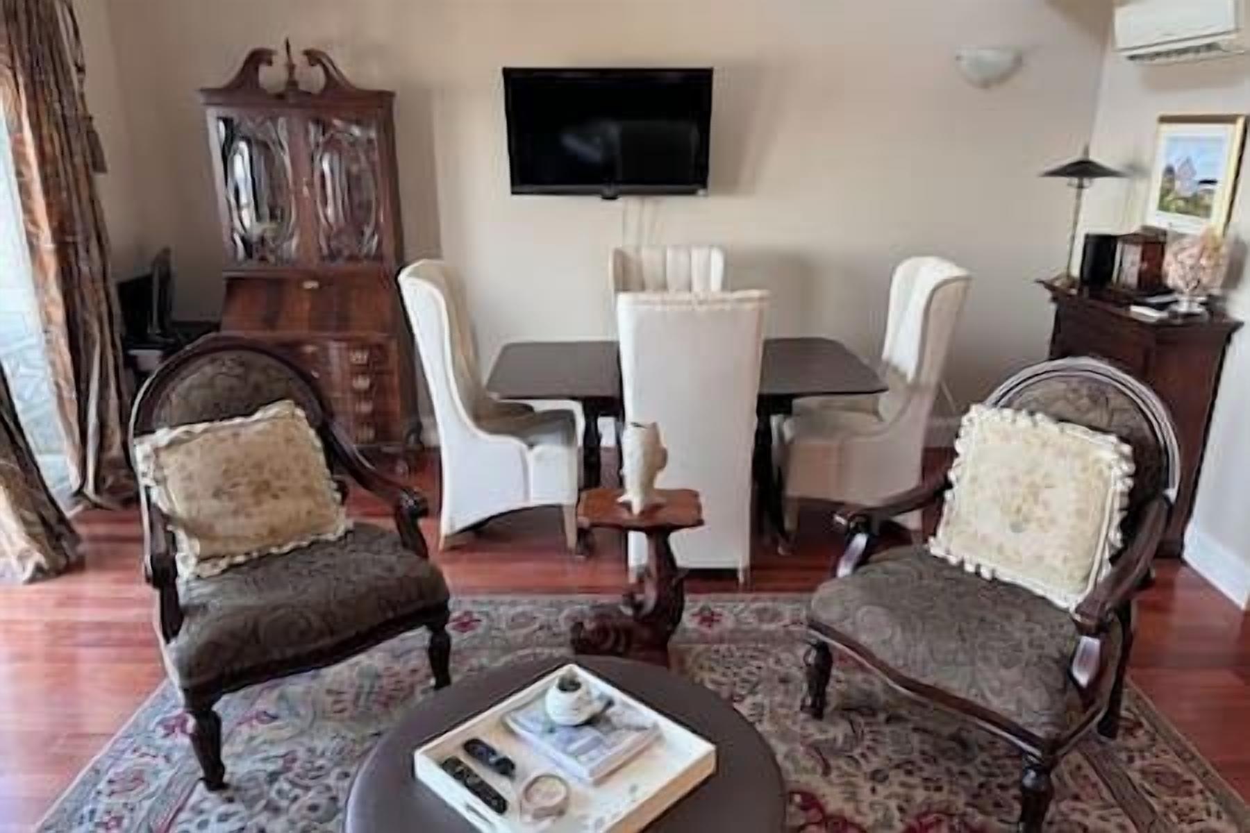 Un salon avec un téléviseur fixé au mur, une table à manger avec des chaises et deux fauteuils avec des coussins.