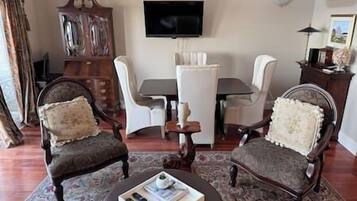 Un salon avec un téléviseur fixé au mur, une table à manger avec des chaises et deux fauteuils avec des coussins.