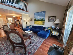 A living room with a blue sofa, a wooden chair, a round ottoman, a rug, a painting, and a kitchen in the background.