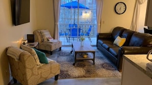 A living room with a sofa, armchair, and a coffee table. There is a dining area with a table and chairs outside a glass door. A clock is on the wall. - Cozy Branson Getaway (Branson)