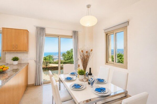 A dining area with a table set for two, a view of the sea, and a kitchen with wooden cabinets.