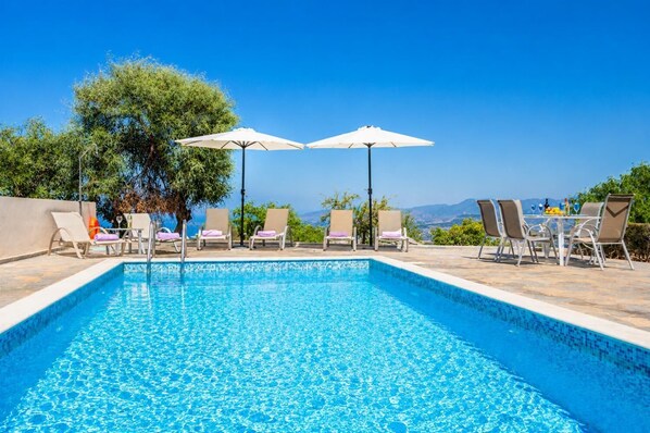 A swimming pool with lounge chairs and umbrellas, surrounded by greenery and a clear view of the mountains.