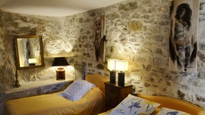 A room with stone walls, two beds with floral bedding, a nightstand with lamps, and artwork on the walls. - Le Castélas (Aigueze)