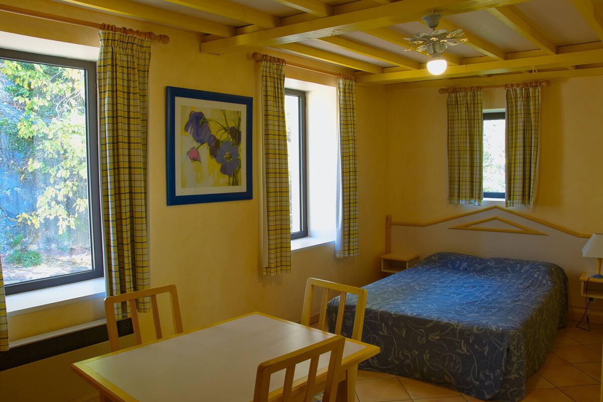 Una habitación con cama, una mesa y sillas. Hay ventanas con cortinas.