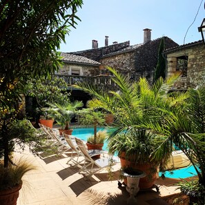 Un área de piscina con sillas de descanso, plantas en macetas y edificios de piedra al fondo.