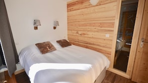 A bedroom with a wooden wall panel, a bed with white bedding, two wall-mounted light fixtures, and a sliding glass door leading to a bathroom. - Ski-In/Ski-Out | Arc 1800 Center | Steps to ESF & Ski Garden (Bourg-Saint-Maurice)