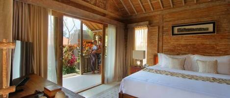 A wooden-floored bedroom with a large bed, a wooden desk with a tray of cups, and a sliding glass door leading to an outdoor area with flowers.