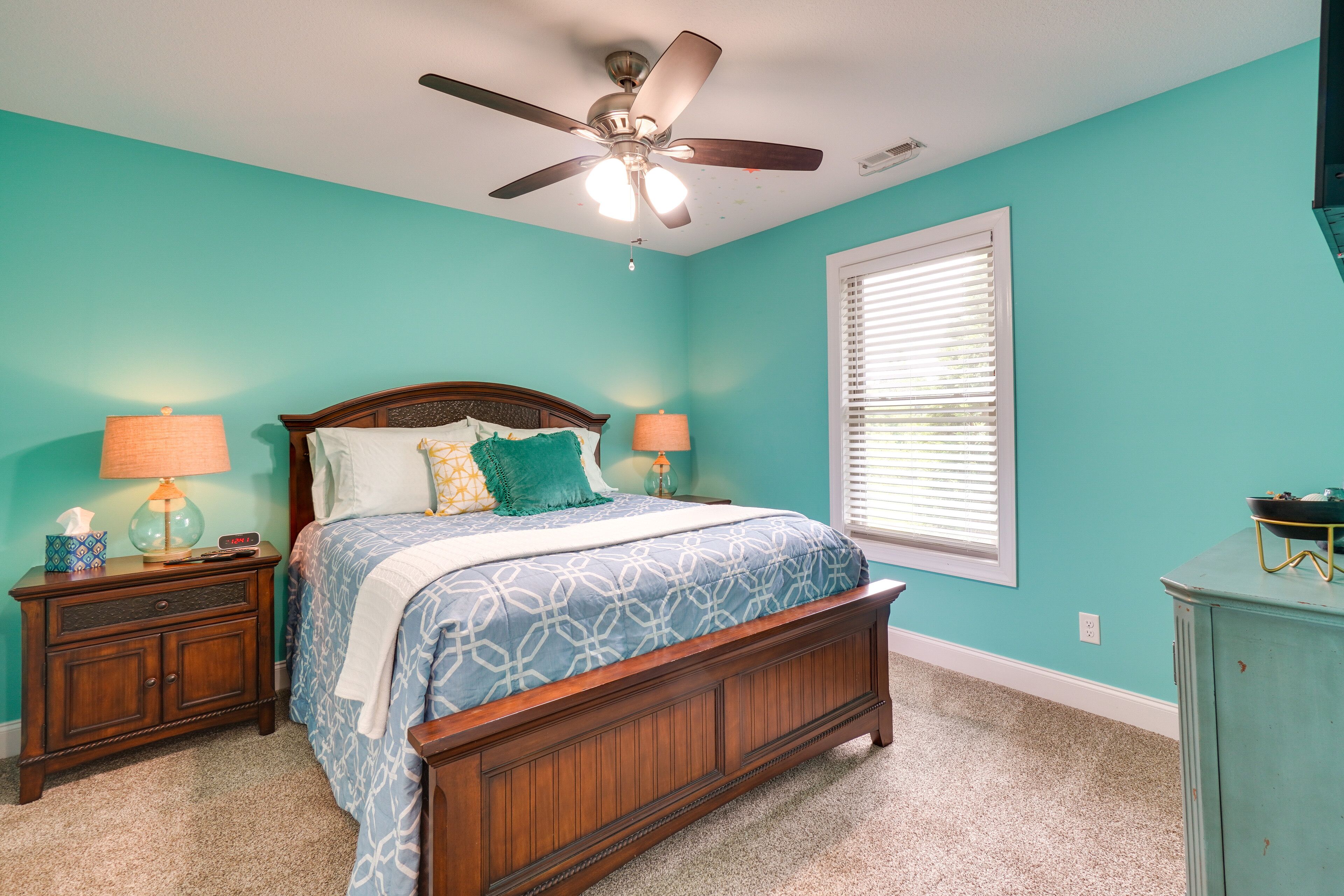 Une chambre à coucher comprenant un lit, deux tables de chevet, un ventilateur de plafond, une fenêtre avec des stores et une commode.