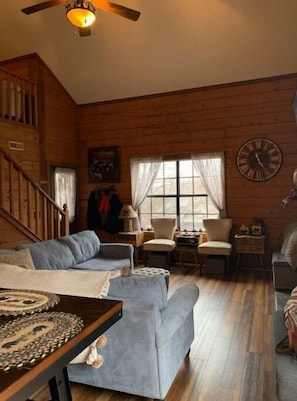 A cozy living room with a wooden interior, a staircase, and a large window with curtains. - Cozy Branson Cabin (Branson)