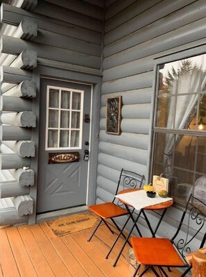 A cabin with a gray exterior, a wooden door with a welcome sign, a window with curtains, and a small table with chairs on the porch. - Cozy Branson Cabin (Branson)
