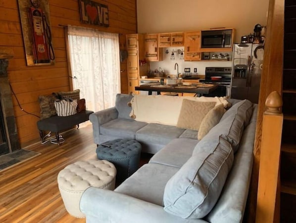 A cozy living room with a grey sectional sofa, a matching ottoman, and a wooden floor. A kitchen area with wooden cabinets and a microwave is visible in the background. - Cozy Branson Cabin (Branson)