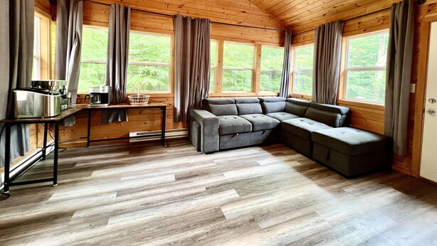 Ein gemütliches Blockhäuschen im Inneren mit einer Eckcouch, einer Kaffeestation und großen Fenstern mit Blick auf den Wald.