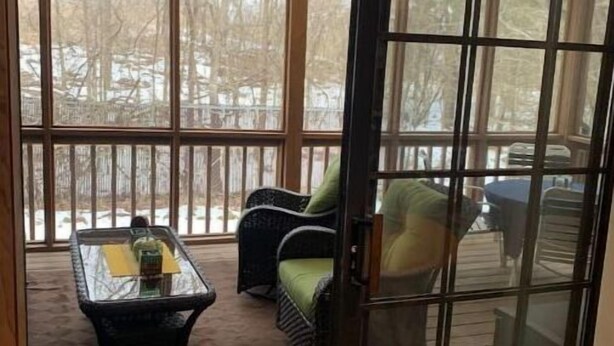 A covered patio area with a glass door, a wicker chair, a small table with a bottle, and a view of a snowy landscape. - Cozy Fall Creek Cabin (Branson)