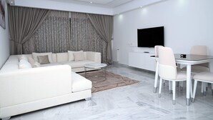 En moderne stue med en hvid sofa, et spisebord med stole, et fladskærms-tv monteret på væggen og en mønstret gardin.