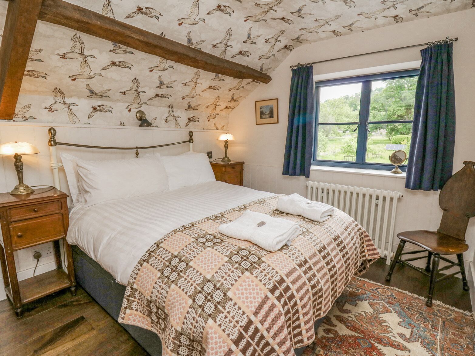 Een slaapkamer met een bed, nachtkastje, stoel, raam met gordijnen en een gestreept plafond.