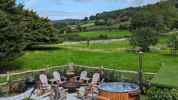 A hot tub surrounded by wooden chairs and a fire pit, set against a scenic hillside backdrop.