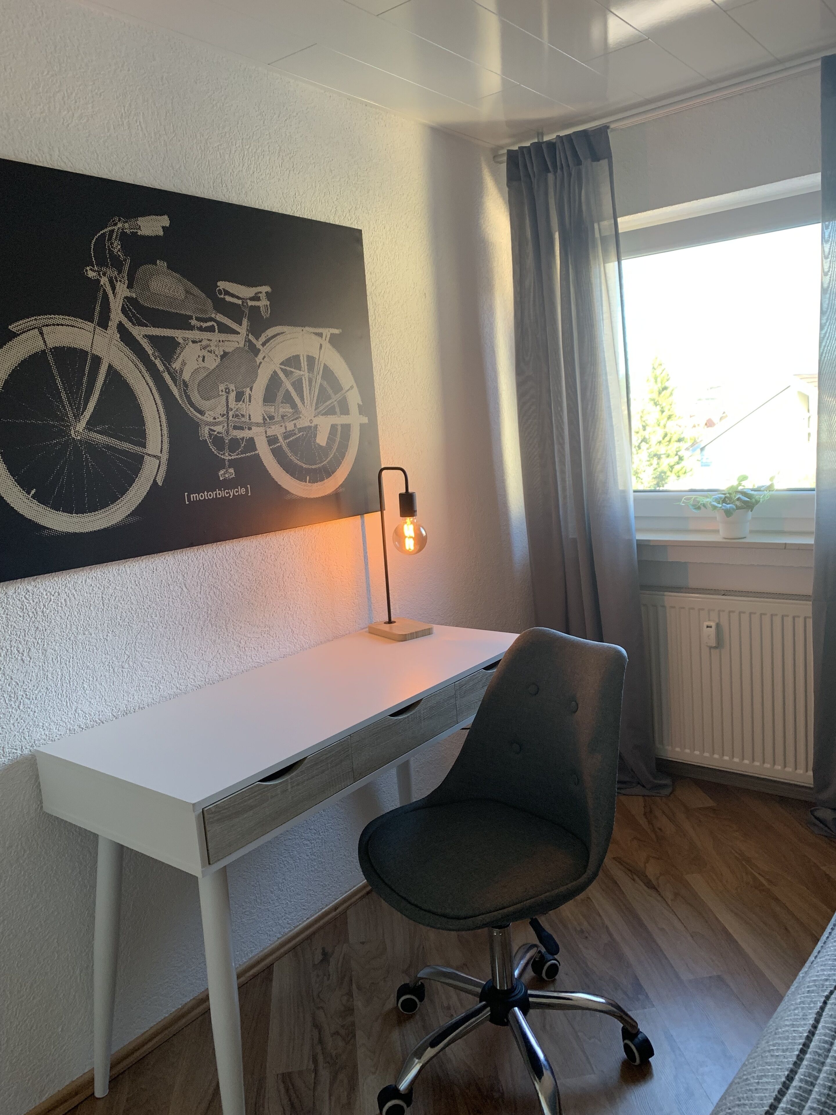 Een moderne kantoorruimte met een wit bureau, een zwarte bureaustoel, een wandlamp en een raam met gordijnen.