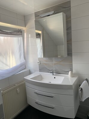 A modern bathroom with a white sink, a mirror above it, and a window with a white curtain. - 2 Zimmer Ferienwohnung (Obersulm)
