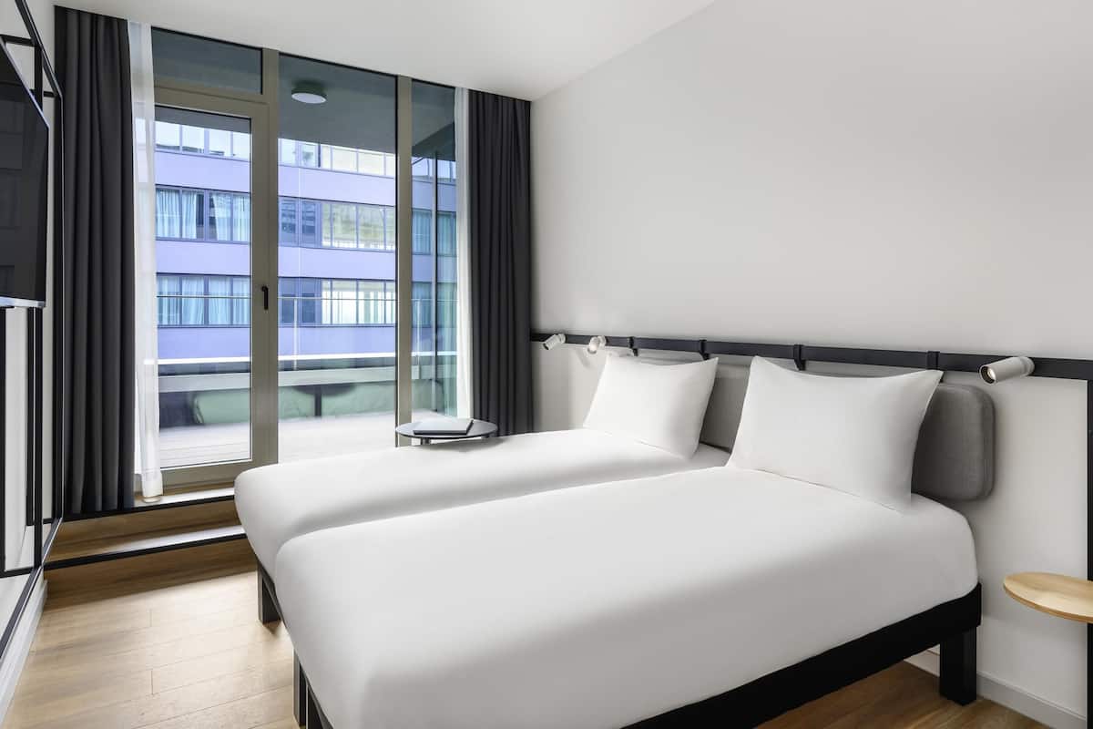 A modern hotel room with a large bed, a small bedside table, and a view of a building through the window.