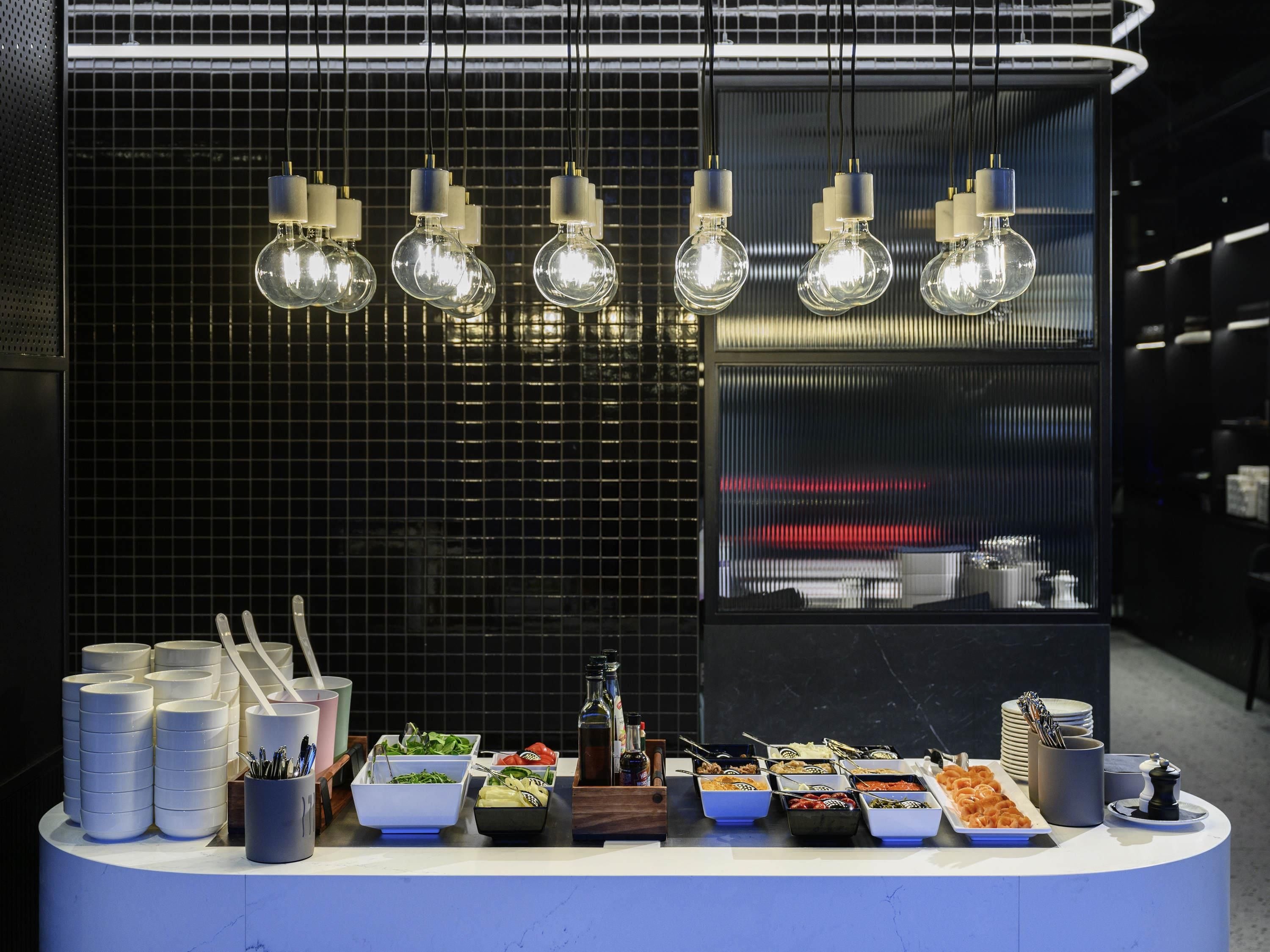 Disposição de buffet com diversos alimentos e utensílios, parede revestida com azulejos e luminárias pendentes.
