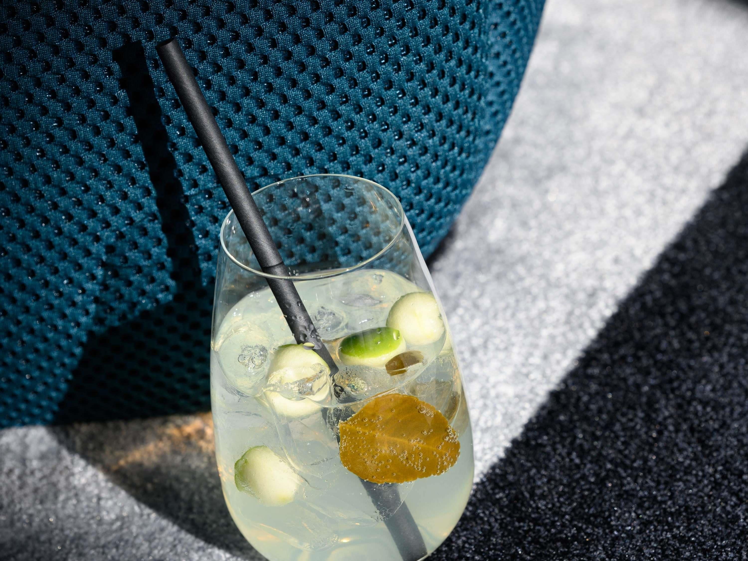 Een glas helder water met ijsblokjes, schijfjes limoen en een geel fruit, mogelijk een citroen, met een zwart rietje.