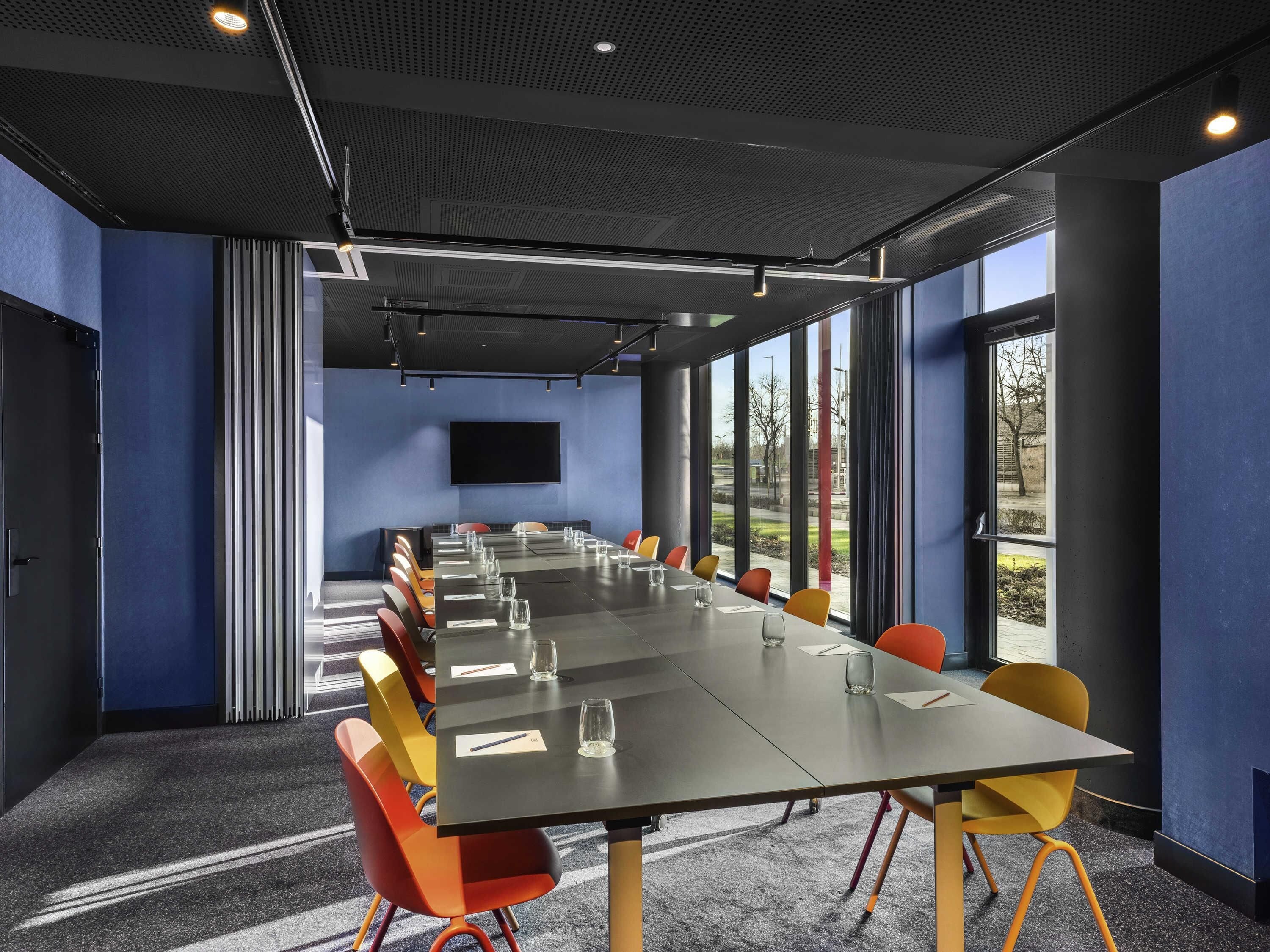 Un salón de conferencias con una mesa larga, sillas, un televisor de pantalla plana y ventanas amplias.