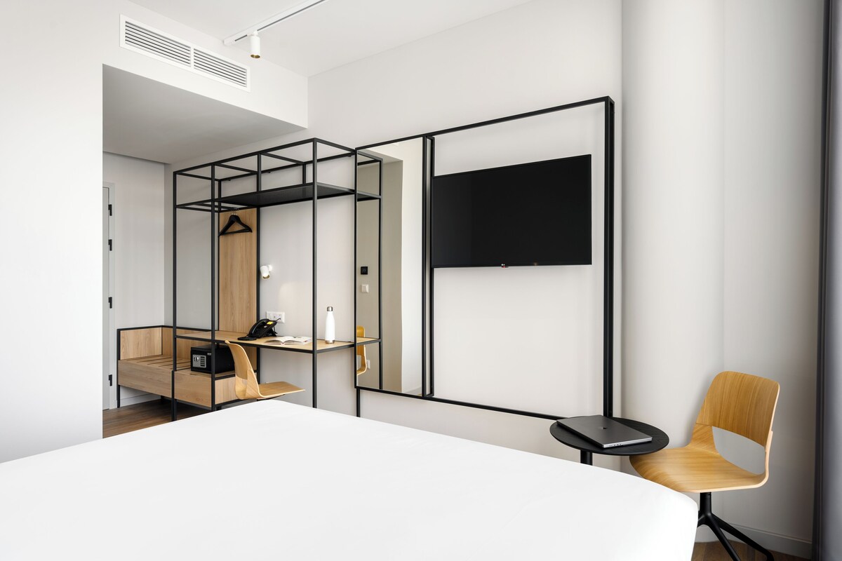 A modern hotel room with a bed, a desk, a chair, a TV, and a wardrobe.