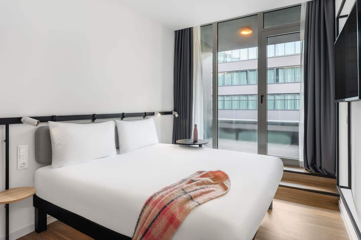 A modern hotel room with a large bed, a bedside table, a nightstand, a window with curtains, and a view of the city.