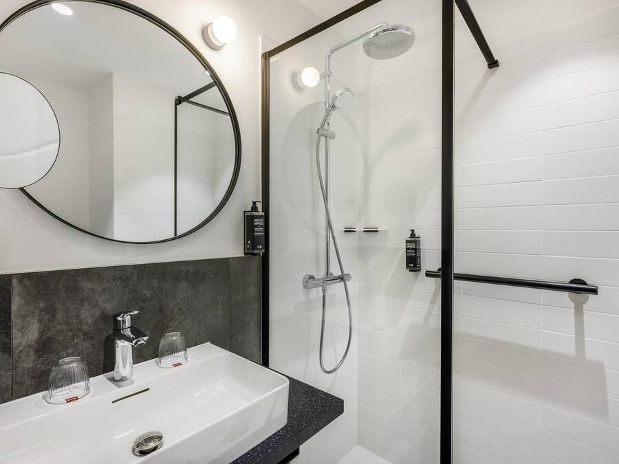 Un baño moderno con un lavabo blanco, un espejo redondo y una zona de ducha con paredes de vidrio.