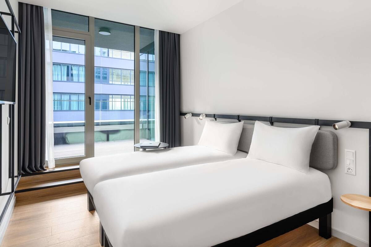 A modern hotel room with a large bed, a small bedside table, a window with curtains, and a view of a building outside.