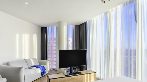 A hotel room with a bed, a TV on a wooden stand, a sofa, and a window with curtains. - ibis Budapest Stadium  (Budapest)