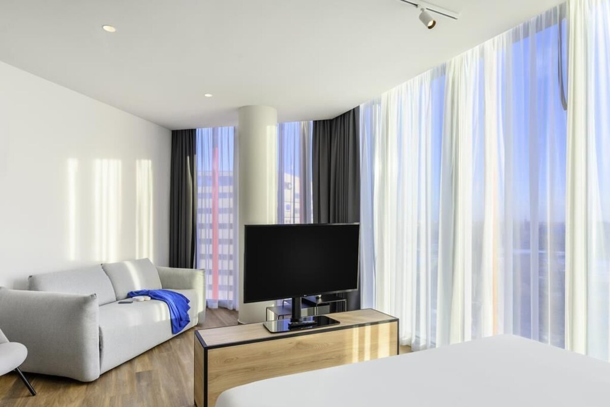 A hotel room with a bed, a TV on a wooden stand, a sofa, and a window with curtains.