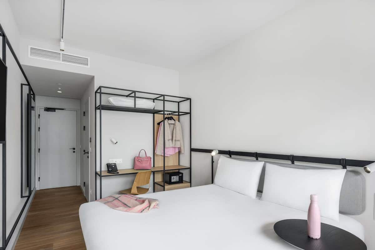 A hotel room with a bed, a desk with a chair, a wardrobe, and a small table with a pink bottle.