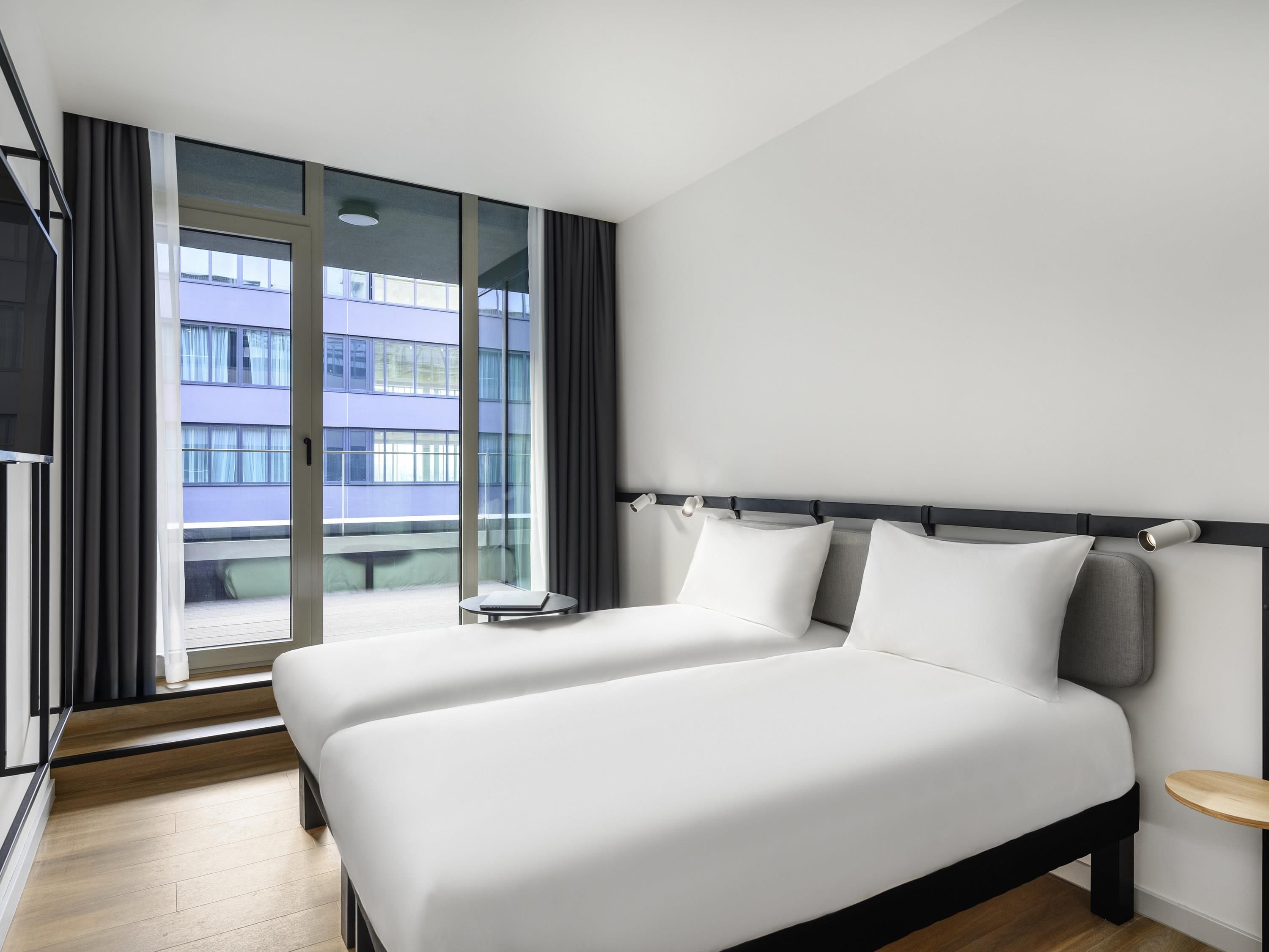 A modern hotel room with a large bed, a small bedside table, and a view of a building through the window.