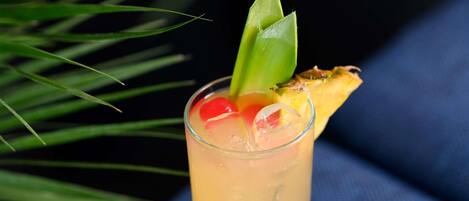 A glass with a tropical drink garnished with a slice of pineapple and a green leaf, set against a dark background with a hint of green foliage.