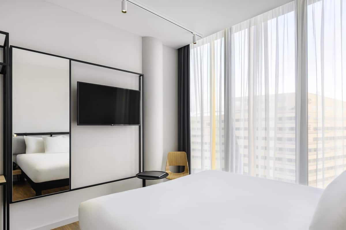 A modern hotel room with a large bed, a flat-screen TV mounted on the wall, a small side table, and a view of the city through sheer curtains.