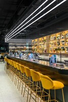 Ένας χώρος μπαρ με κίτρινα σκαμπό, έναν μακρύ πάγκο και ράφια γεμάτα με μπουκάλια και ποτήρια.