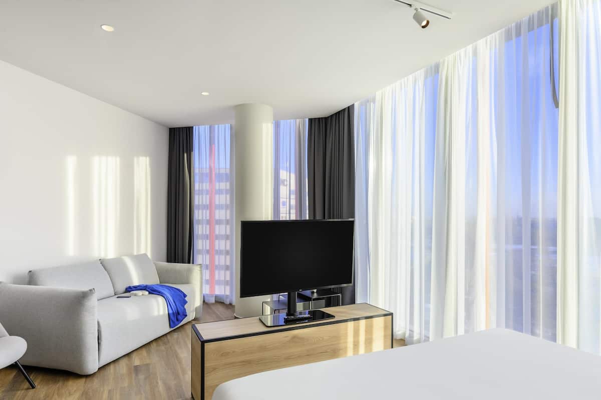 A modern hotel room with a bed, a sofa, a TV, and a desk.