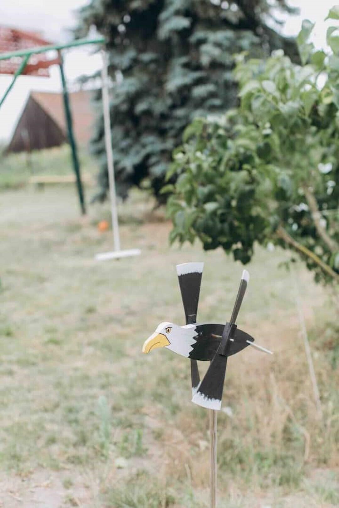 Eine vogelartige Dekoration mit gelbem Schnabel und schwarz-weißen Flügeln, stehend auf einem Pfahl in einer grünen Umgebung mit Bäumen und einem Gebäude im Hintergrund.